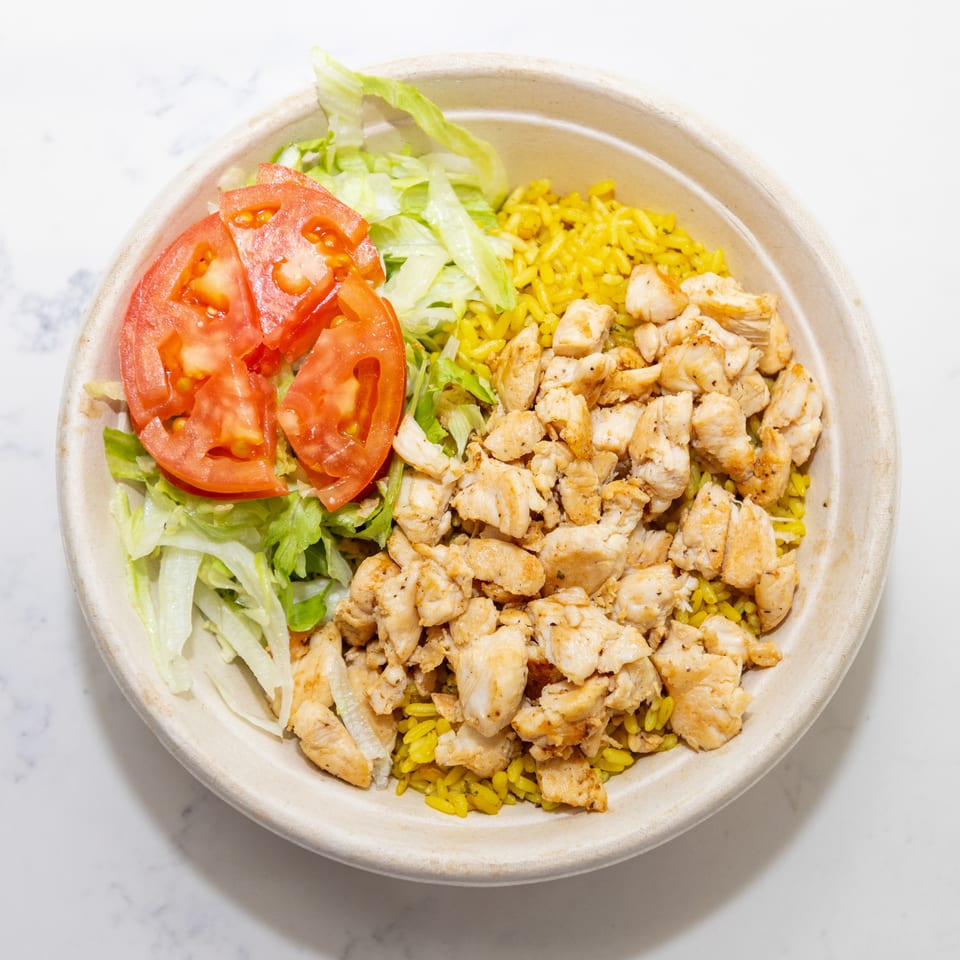 Bowl of seasoned yellow rice topped with grilled chicken, shredded lettuce, and sliced tomatoes on a marble surface.