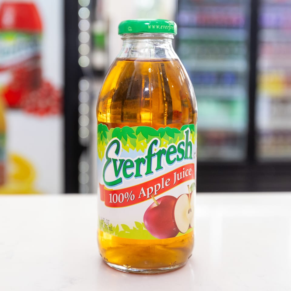 Bottle of VertiFresh 100% apple juice displayed on a white counter, with colorful beverage bottles blurred in the background.