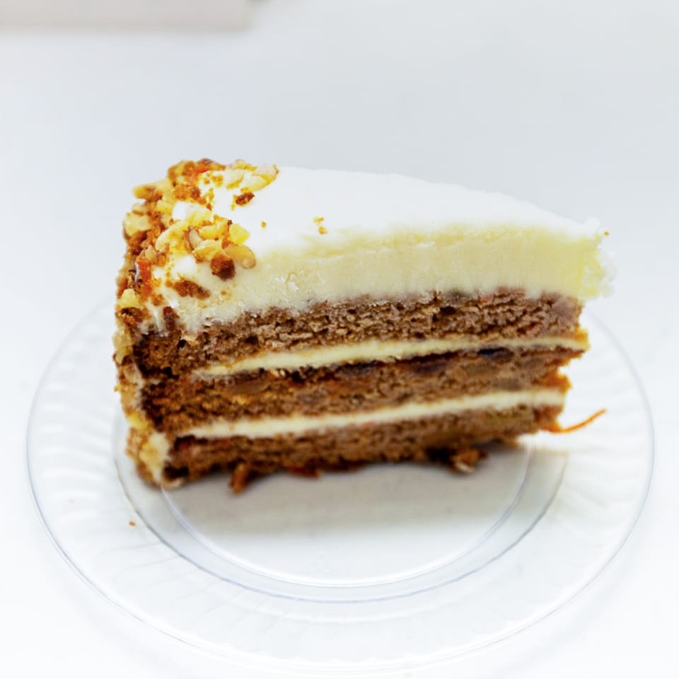 Slice of layered chocolate cake with white frosting and nut topping on a clear glass plate.
