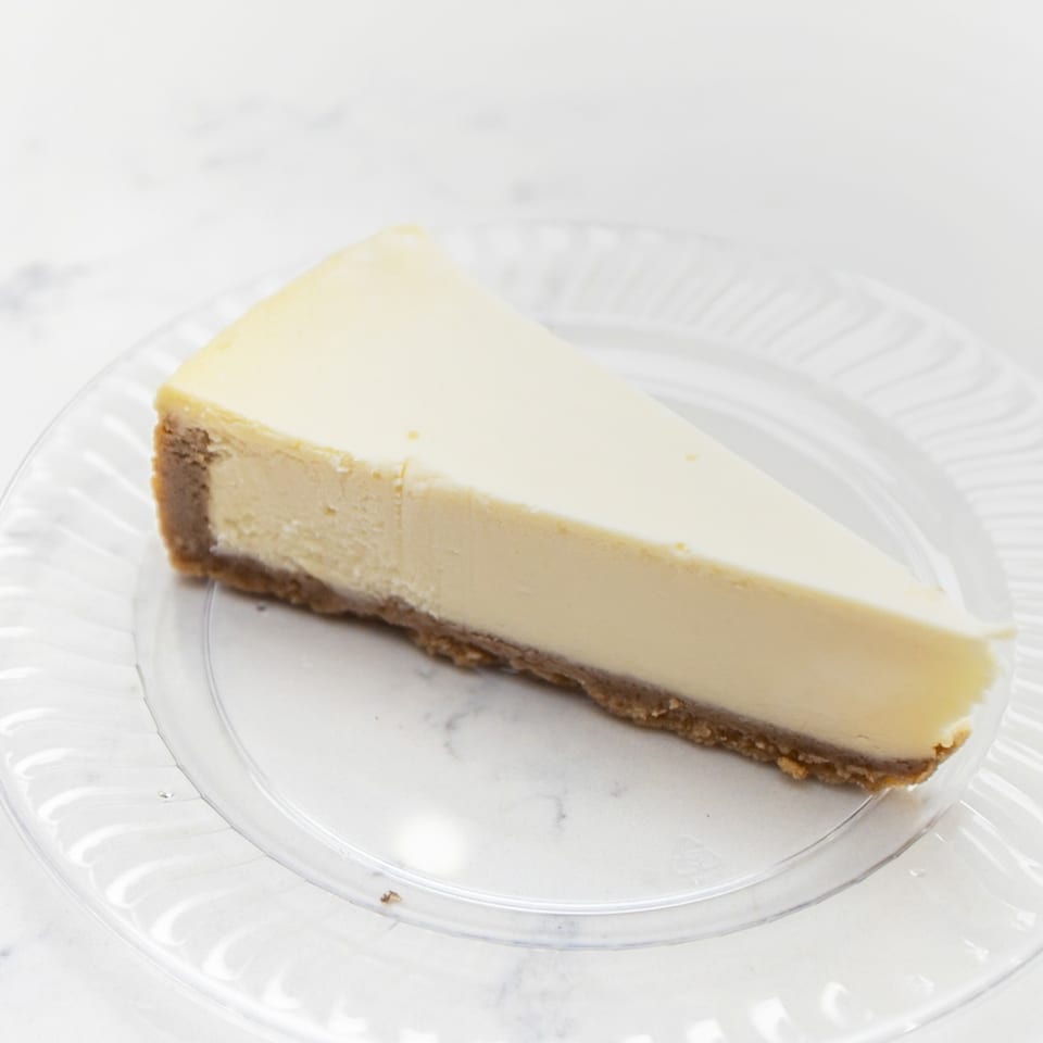 Slice of creamy cheesecake with a graham cracker crust on a clear glass plate.